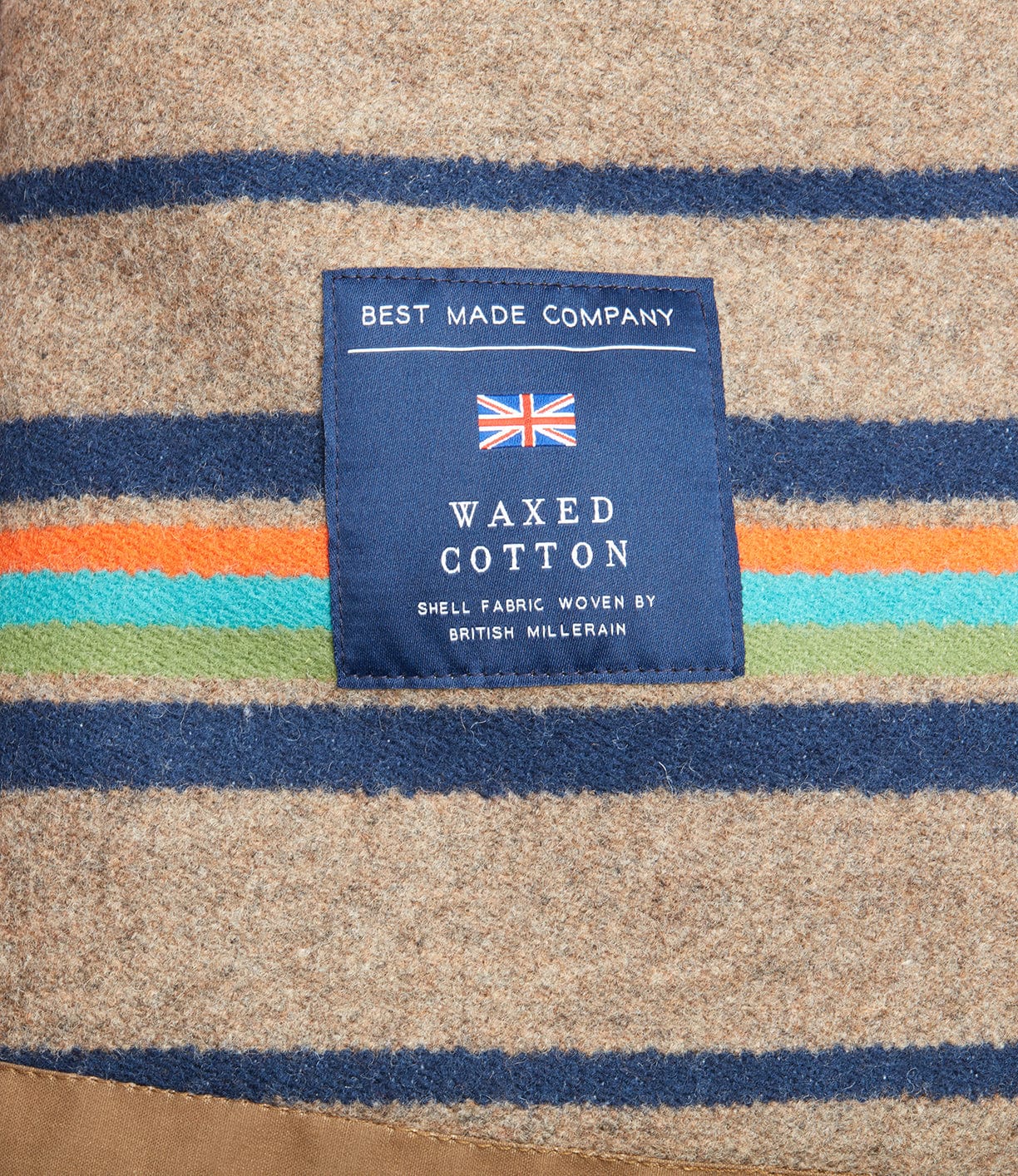 A blue fabric label with a British flag reading Best Made Co. is sewn onto tan wool-lined fabric with blue, orange, and green stripes—perfect for the Wool-lined Waxed Field Jacket Tan.