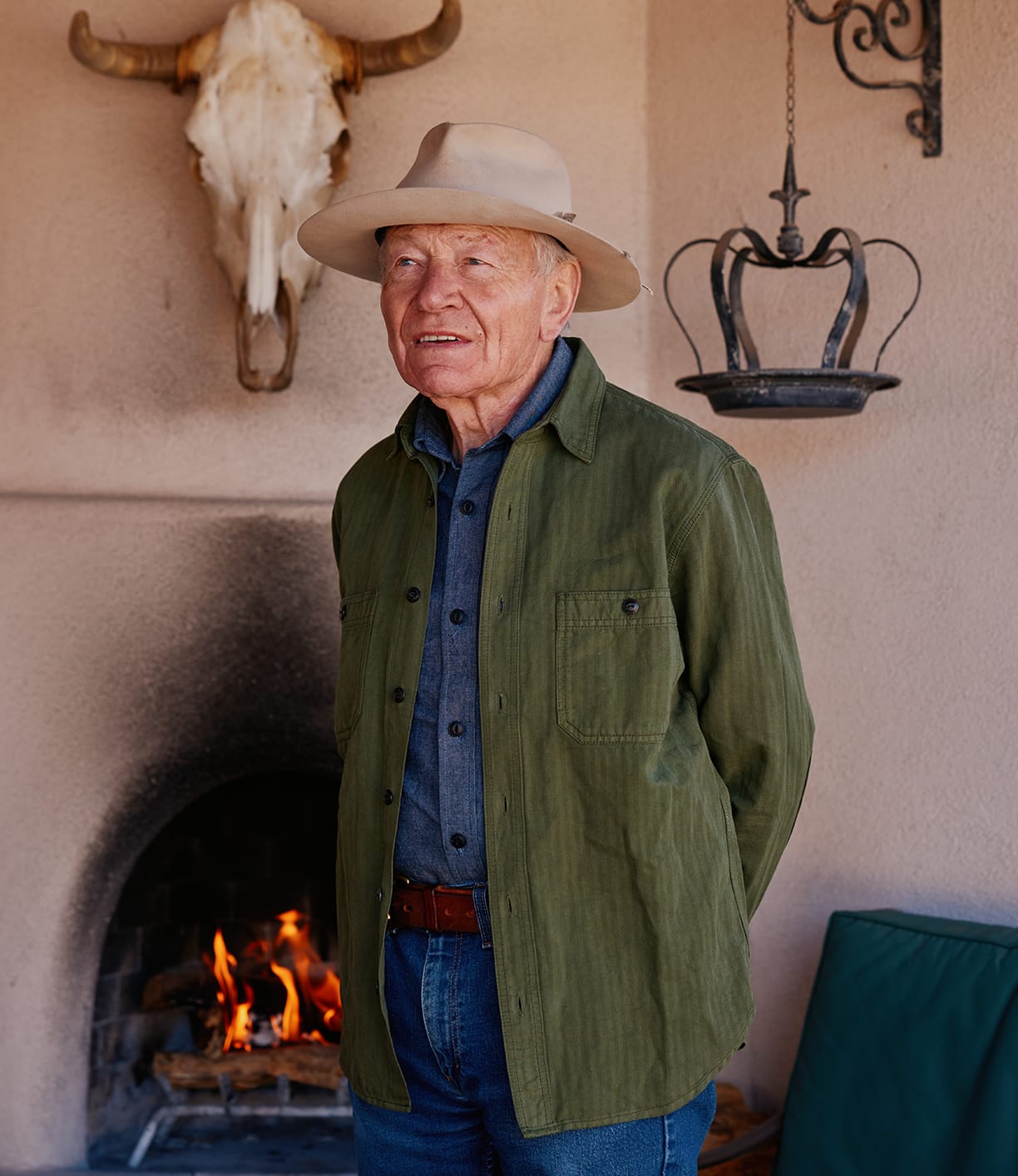 An older man in a hat and green jacket stands before a lit fireplace, wearing the Best Made Co. Cotton-Linen Workshirt in Olive Drab, bringing rugged charm to the rustic setting with a cow skull above and a metal lantern nearby.