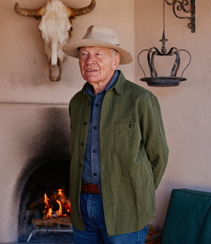 An older man in a hat and green jacket stands before a lit fireplace, wearing the Best Made Co. Cotton-Linen Workshirt in Olive Drab, bringing rugged charm to the rustic setting with a cow skull above and a metal lantern nearby.