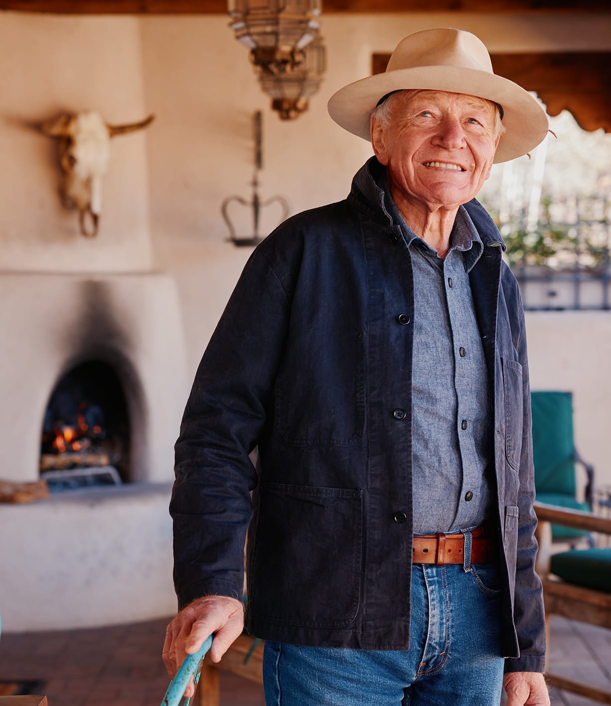 An older man in a light wide-brim hat, blue shirt, and Best Made Co. Service Jacket Special Edition Navy smiles on a porch with a lit fireplace and a bull skull mounted on the wall in the background.