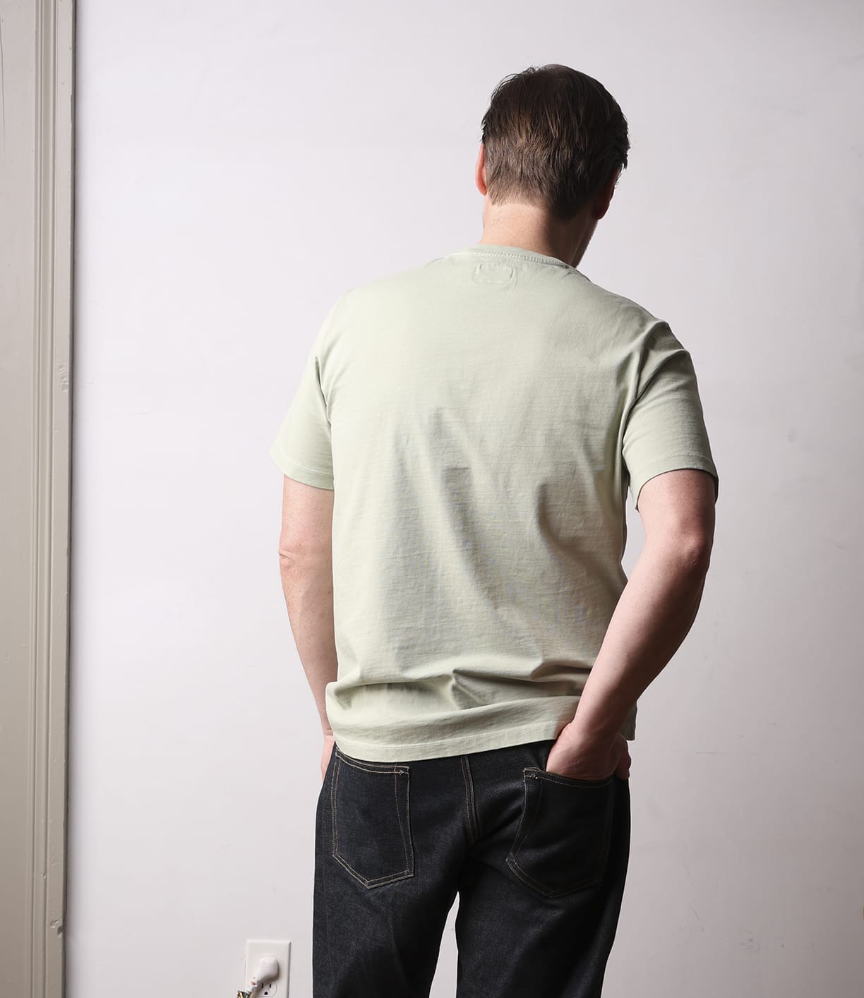 A person stands against a light wall wearing the Best Made Co. Standard Pocket T-Shirt Haze—crafted from durable Peruvian tangüis cotton—with dark jeans. One hand is in their pocket, the other on their lower back, highlighting this everyday staple.