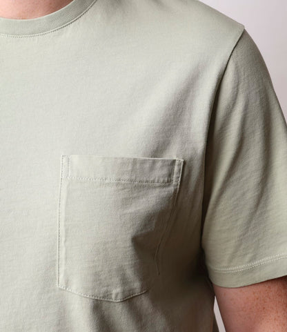 Close-up of someone wearing the Best Made Co. Standard Pocket T-Shirt in Haze, a light green, garment-dyed Peruvian tangüis cotton tee with a left chest pocket. The photo shows the upper torso and part of an arm, not the face.
