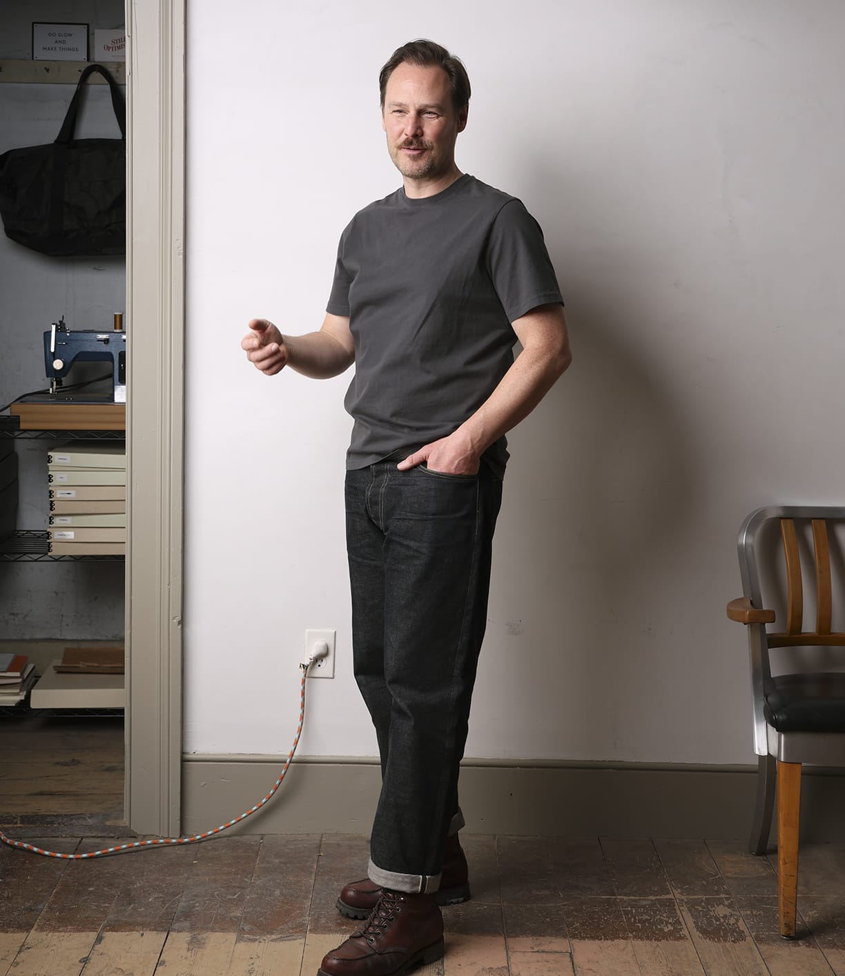 A man indoors stands on a wooden floor near a wall socket, wearing the Best Made Co Standard T-Shirt in Faded Black with dark jeans and brown boots. One hand in his pocket, he gestures; a chair and sewing machine complete this everyday staple look.