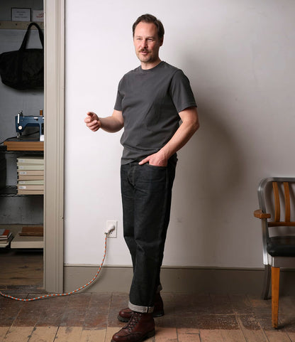 A man wearing the Best Made Co. Standard T-Shirt in Faded Black and dark jeans stands indoors on a wooden floor, one hand in his pocket and the other gesturing, with a chair, bookshelves, and a vintage sewing machine behind him.