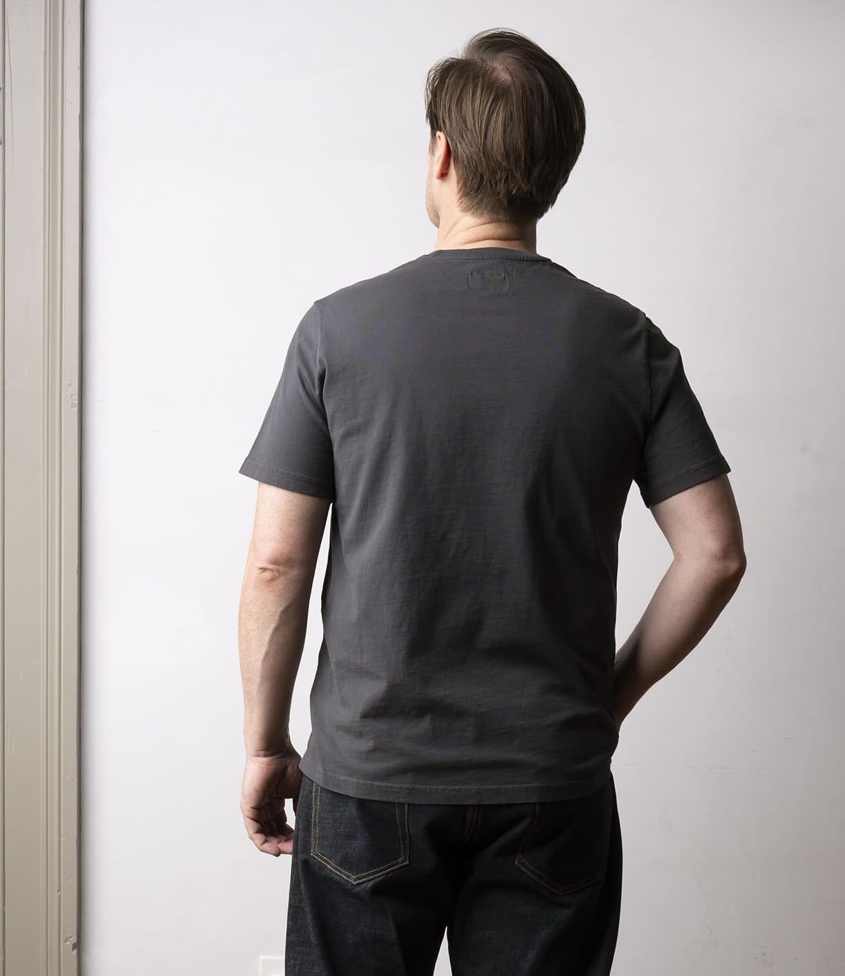 A man faces away from the camera, wearing Best Made Co’s Standard T-Shirt in Faded Black with dark jeans—a versatile essential—standing against a plain light-colored wall.