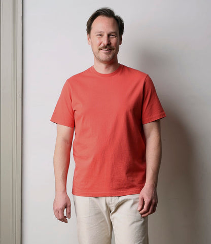 A man with short brown hair and a mustache stands smiling against a plain light wall, wearing the Best Made Co. Standard T-Shirt in faded red—an everyday essential, paired with light beige pants.