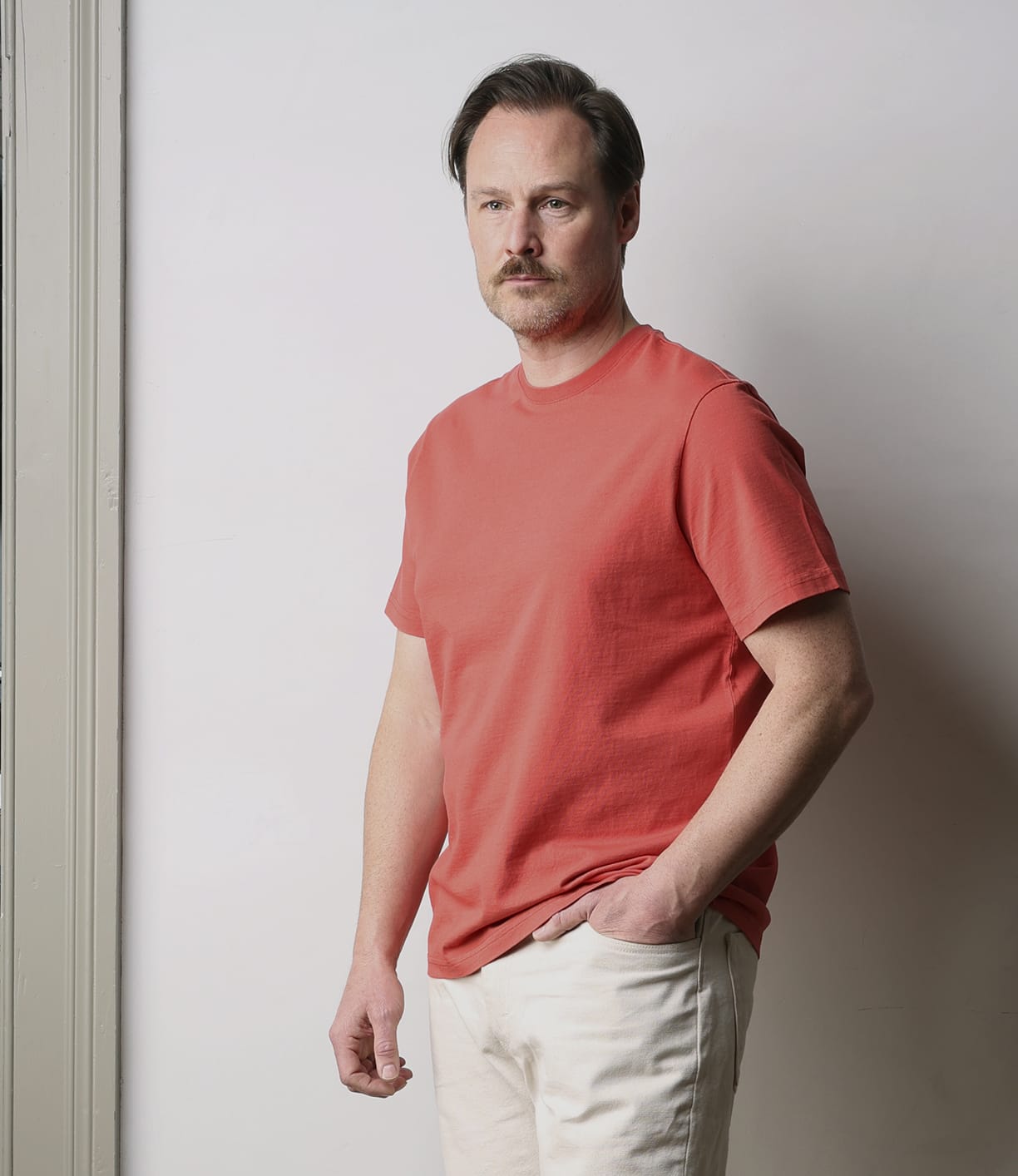 A man with short brown hair and a mustache wears the Best Made Co. Standard T-Shirt in Faded Red and light-colored pants, standing indoors against a plain light gray wall with one hand in his pocket.