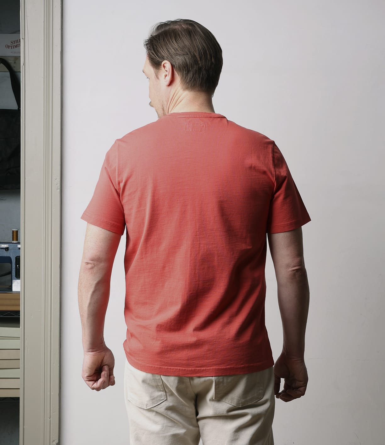 A man with short brown hair, wearing the Best Made Co. Standard T-Shirt in Faded Red and beige pants, stands indoors facing away from the camera, highlighting the back of his everyday staple outfit.