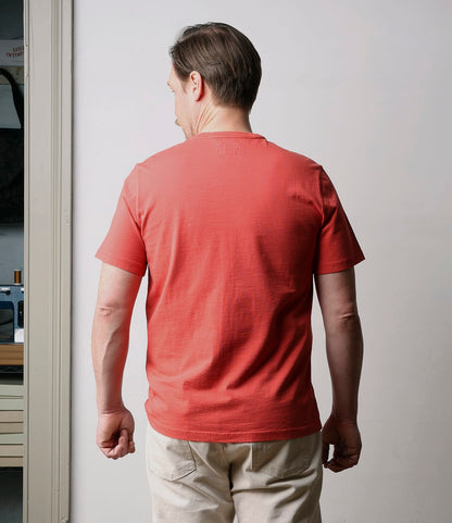 A man with short brown hair, wearing the Best Made Co. Standard T-Shirt in Faded Red and beige pants, stands indoors facing away from the camera, highlighting the back of his everyday staple outfit.