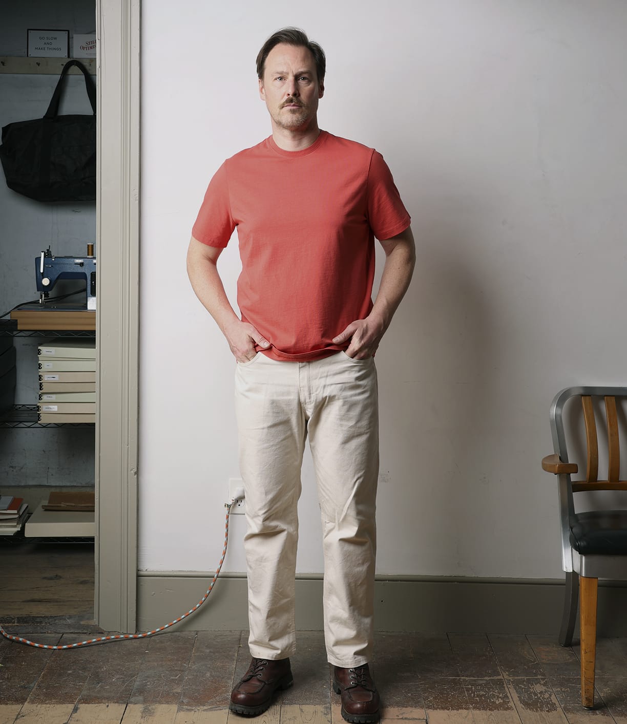 A man stands indoors by bookshelves, wearing Best Made Co.'s Standard T-Shirt in Faded Red—crafted from soft Peruvian tangüis cotton—paired with beige pants and brown shoes. Nearby are books, a sewing machine, and a wooden chair.