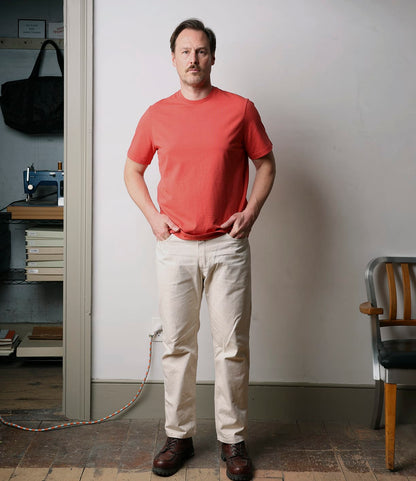 A man stands indoors by bookshelves, wearing Best Made Co.'s Standard T-Shirt in Faded Red—crafted from soft Peruvian tangüis cotton—paired with beige pants and brown shoes. Nearby are books, a sewing machine, and a wooden chair.