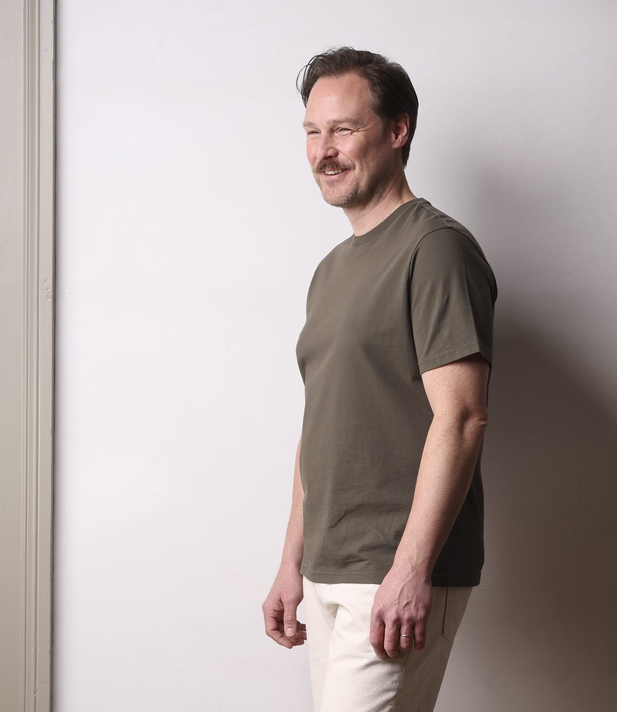A smiling man with short hair and a mustache stands in profile against a light wall, wearing Best Made Co.'s Standard T-Shirt Field Drab and light pants, his arms relaxed by his sides.