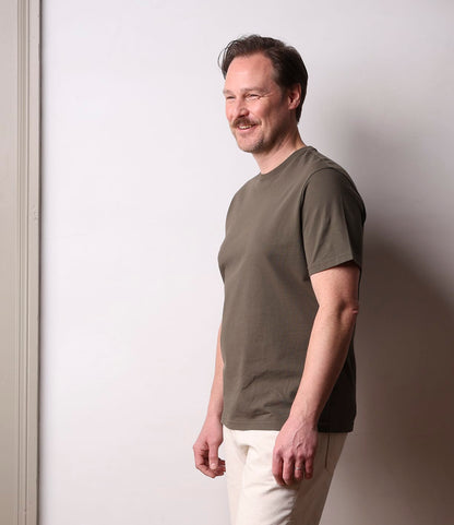A smiling man with short hair and a mustache stands in profile against a light wall, wearing Best Made Co.'s Standard T-Shirt Field Drab and light pants, his arms relaxed by his sides.