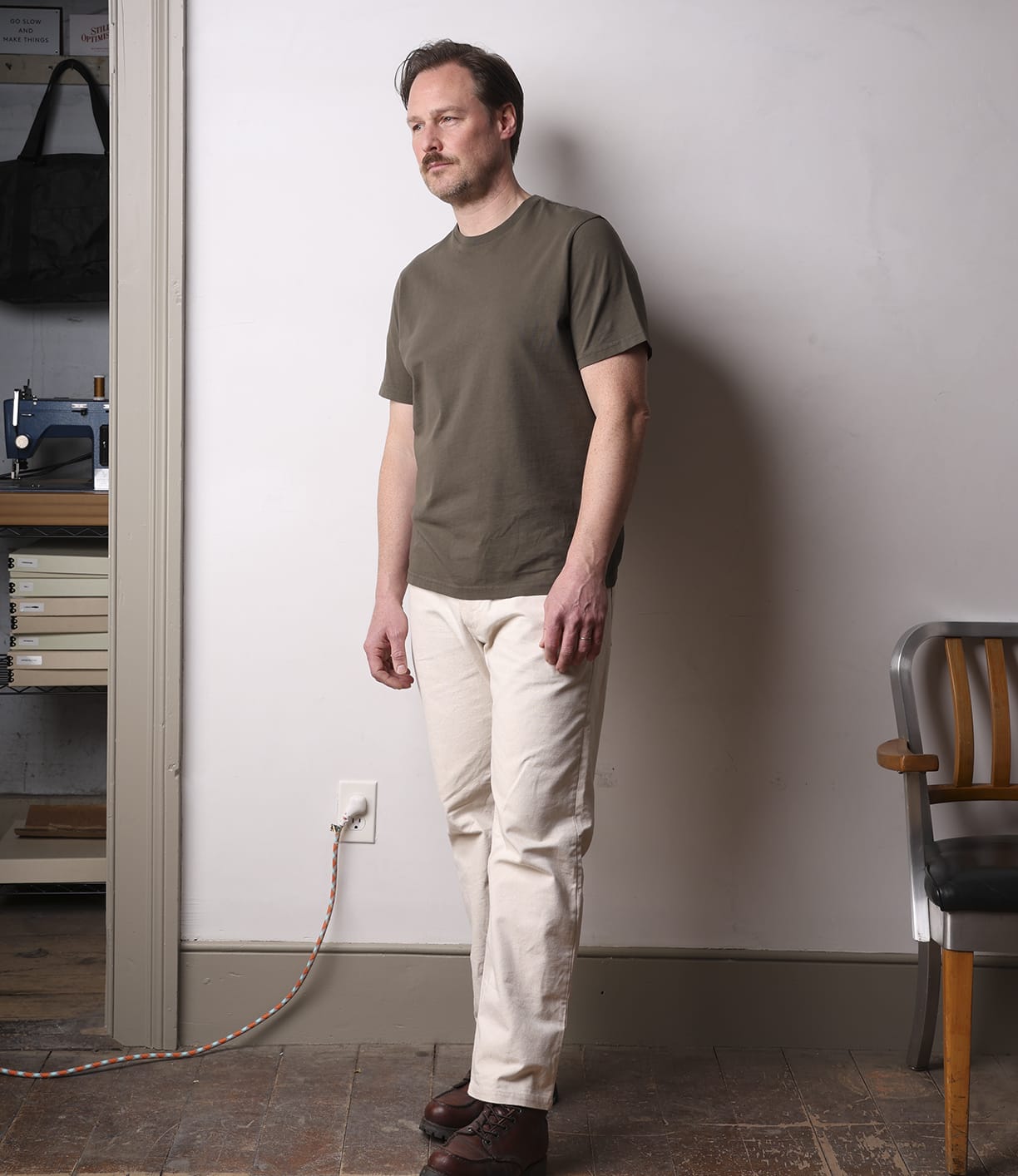 A man stands indoors against a white wall, wearing the Best Made Co. Standard T-Shirt in Field Drab, light-colored pants, and brown shoes. Next to him is a wooden chair and a nearby plugged-in power cord.