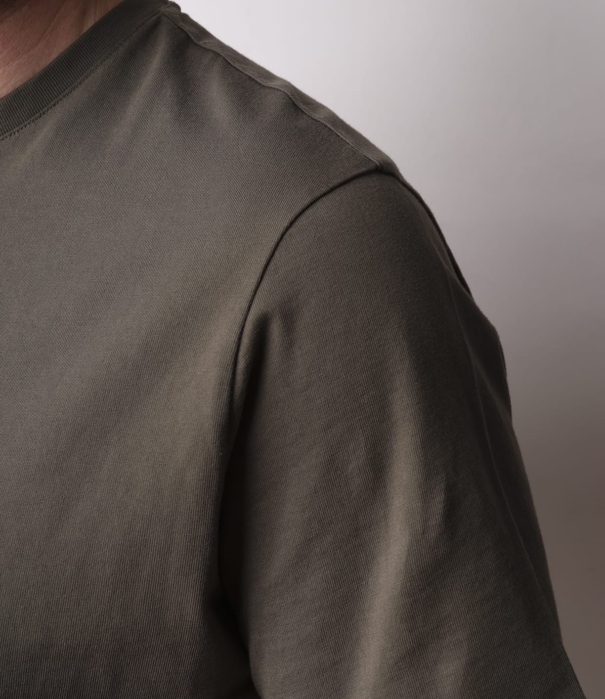 Close-up of a person wearing the Best Made Co. Standard T-Shirt Field Drab, featuring soft Peruvian tangüis cotton in olive green. Only the shoulder, upper chest, and sleeve are visible against a plain light background; head and lower body are out of frame.
