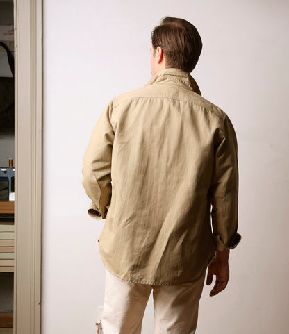 A man stands indoors near a doorway, wearing the Best Made Co. Cotton-Linen Workshirt Tan and off-white pants, with shelves and a sewing machine visible in the next room.