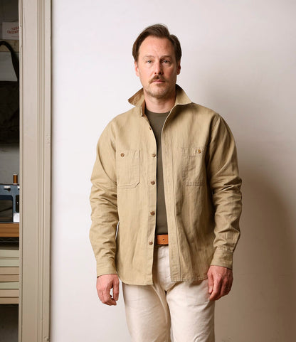 A man with light brown hair and a mustache stands indoors against a plain wall, wearing the Best Made Co. Cotton-Linen Workshirt Tan over a dark undershirt and light pants, looking directly at the camera.