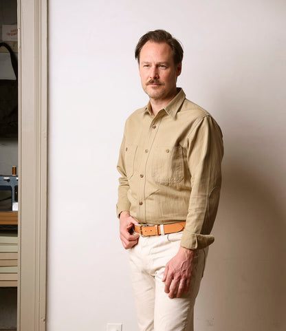 A man with light brown hair and a mustache stands indoors against a plain wall, wearing the Best Made Co. Cotton-Linen Workshirt in tan, paired with light pants and a tan belt, looking at the camera with a neutral expression.