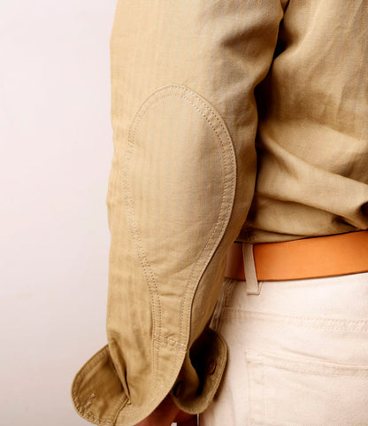 Close-up of a person wearing the Best Made Co. Cotton-Linen Workshirt in Tan, featuring an oval elbow patch and paired with cream pants and a tan belt. The sleeve is slightly wrinkled. Made in Portugal.