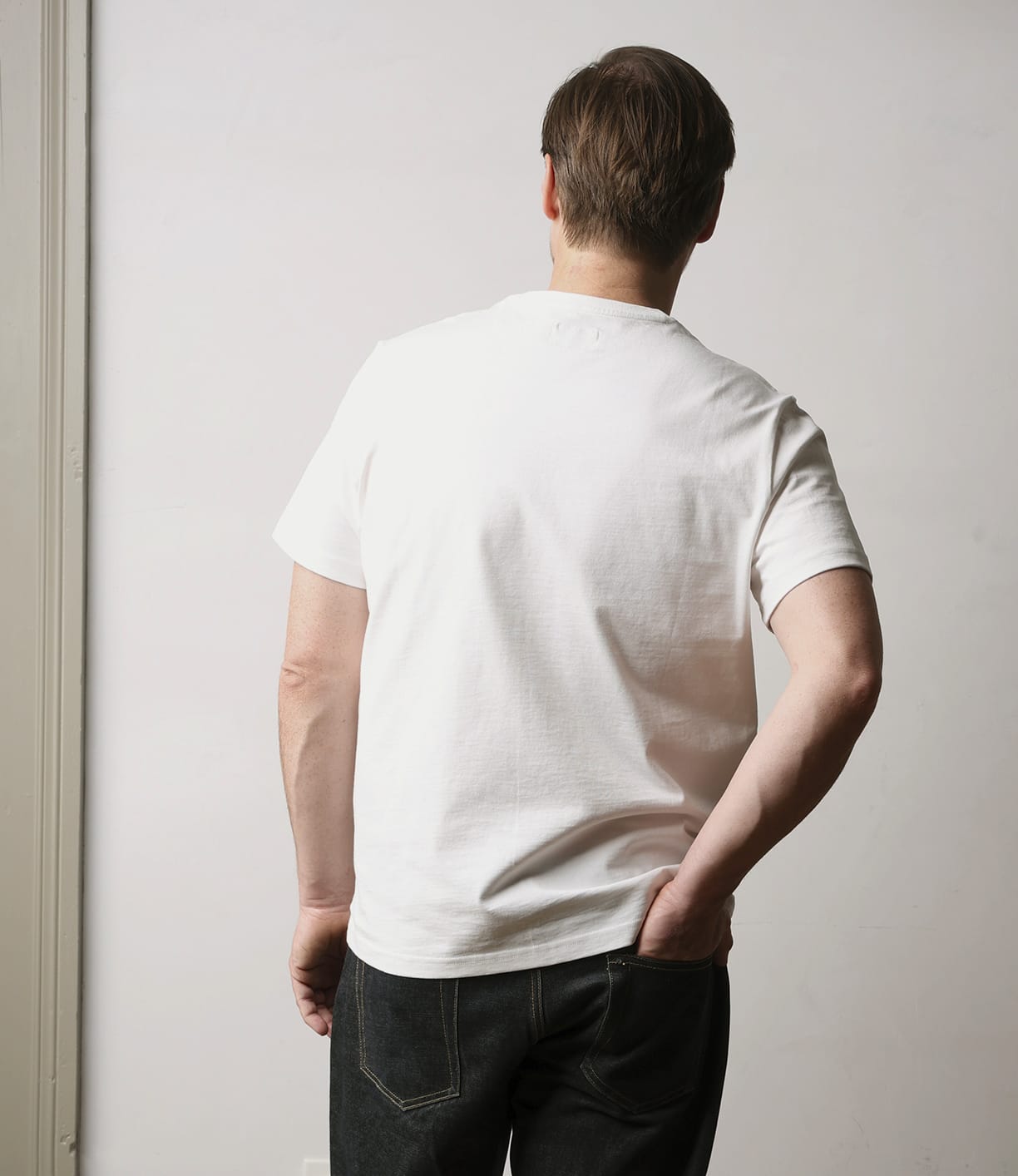A person with short brown hair, wearing the Best Made Co. Standard T-Shirt Undyed and dark jeans, stands facing away from the camera with one hand on their hip against a light-colored background.