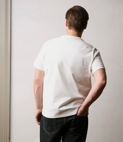 A person with short brown hair, wearing the Best Made Co. Standard T-Shirt Undyed and dark jeans, stands facing away from the camera with one hand on their hip against a light-colored background.
