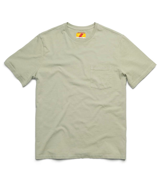 A man with short hair wears the Best Made Co Standard Pocket T-Shirt Haze, paired with dark jeans and brown boots. He stands indoors near a wooden chair and wall outlet, smiling slightly with his hands loosely clasped.