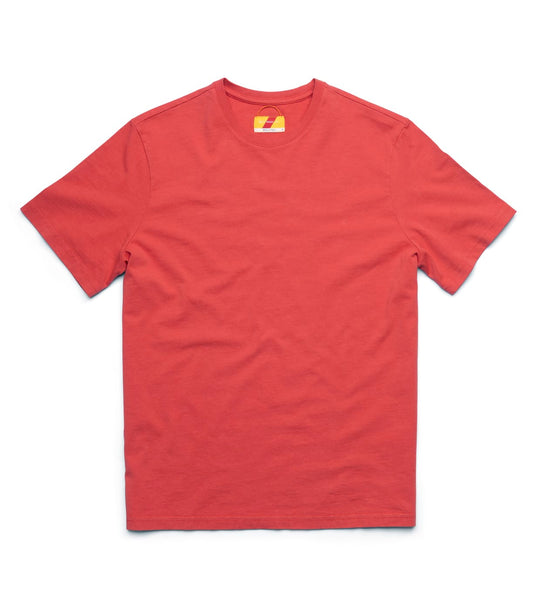 A man stands indoors against a plain wall, wearing the Best Made Co Standard T-Shirt in Faded Red, beige pants, and brown shoes. His hands are in his pockets. A chair, books, and a sewing machine are visible in the background.