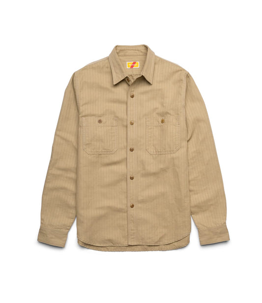 A man with light brown hair and a mustache stands indoors against a plain wall, wearing the Best Made Co. Cotton-Linen Workshirt in tan, paired with light pants and a tan belt, looking at the camera with a neutral expression.