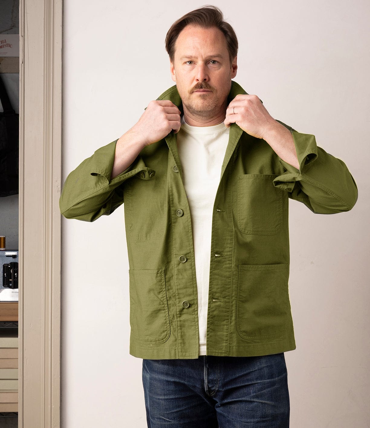 A man with light brown hair and a mustache adjusts the collar of his green Best Made Co. Panama Cloth Service Jacket, inspired by the classic P-41 Marine Corps jacket, while indoors in a white shirt and blue jeans.