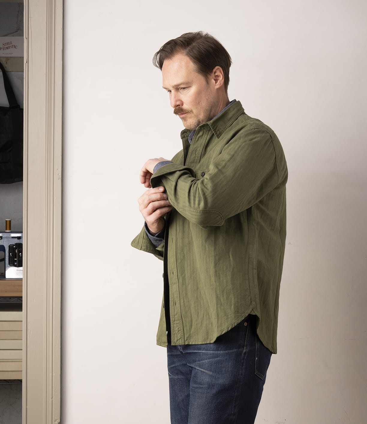 A man with light brown hair stands indoors near a kitchen doorway, looking down while rolling up his sleeve. He is wearing the Best Made Co. Cotton-Linen Workshirt in Olive Drab and blue jeans.