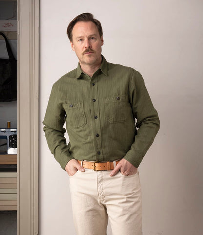 A man with light brown hair and a mustache stands indoors wearing the Best Made Co. Cotton-Linen Workshirt in Olive Drab, beige pants, and a tan belt. He has his hands in his pockets and looks at the camera with a neutral expression.