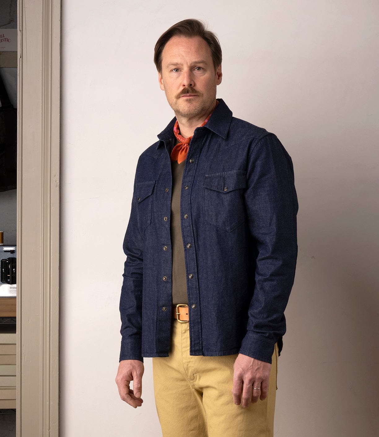 A man with light brown hair and a mustache, wearing the Best Made Co Fukuyama Snap-Front Shirt in indigo denim, tan pants, an olive shirt, and an orange scarf, stands indoors with a neutral expression.