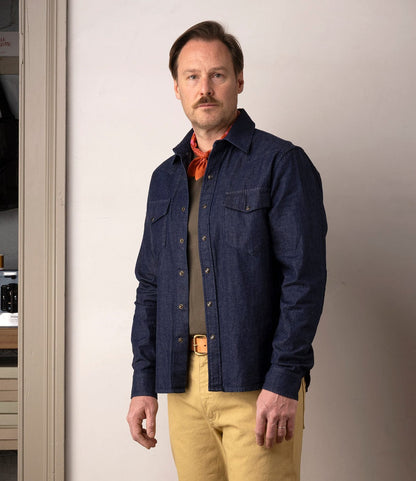 A man with light brown hair and a mustache stands indoors, wearing the Best Made Co. Fukuyama Snap-Front Shirt in Japanese indigo denim, paired with khaki pants, a brown belt, and an orange scarf, looking directly at the camera.