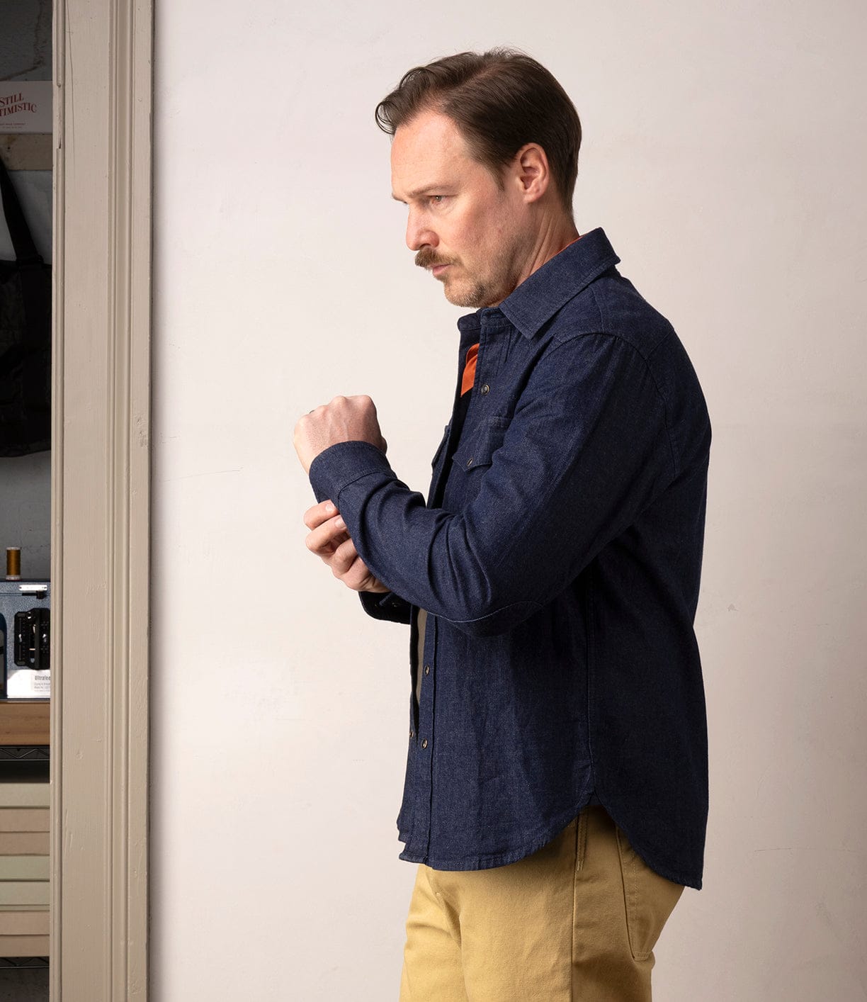 A man with light brown hair and a mustache stands in profile indoors, adjusting his sleeve while wearing the Best Made Co Fukuyama Snap-Front Shirt in indigo with beige pants.