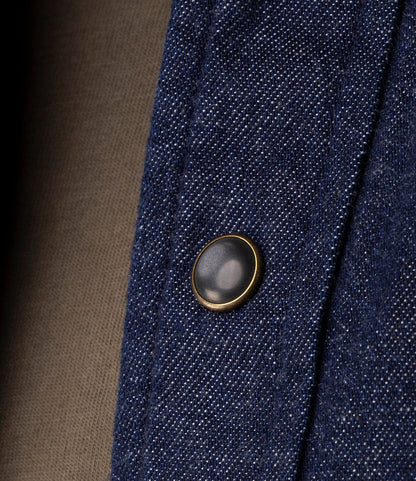 A close-up of Japanese indigo denim on the Best Made Co. Fukuyama Snap-Front Shirt features a metallic snap-front button and visible stitching, with a brown fabric background highlighting the shirt’s western details.
