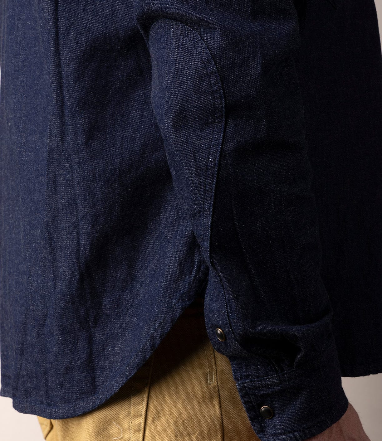 A close-up side view shows a person wearing the Best Made Co Fukuyama Snap-Front Shirt—an indigo-dyed Japanese denim shirt with buttoned cuffs—paired with tan pants, the curved hem draping over their relaxed arm.