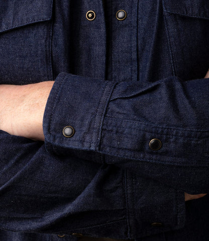 A person wears a dark blue Best Made Co. Fukuyama Snap-Front Shirt with arms crossed, showcasing the sleeve details and signature metal snaps on this Japanese indigo denim snap-front shirt.
