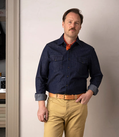 A man with light brown hair and a mustache stands indoors against a plain wall, wearing the Best Made Co. Fukuyama Snap-Front Shirt in Japanese indigo denim, light tan pants, a tan belt, and a red neckerchief, with one hand in his pocket.