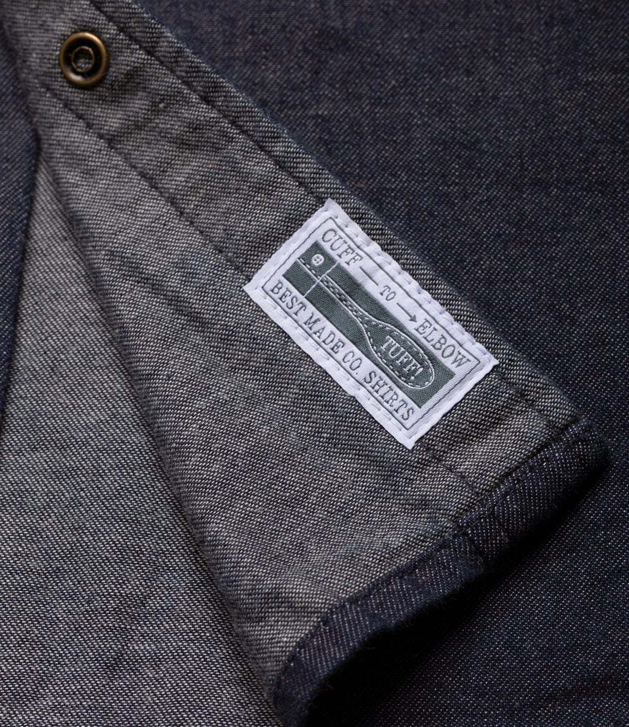 A close-up of the inside cuff of the dark Fukuyama Snap-Front Shirt by Best Made Co shows a white label with green and black text, “CUFF TUFF, BEST MADE CO SHIRTS,” and a brass-colored button on the cuff.