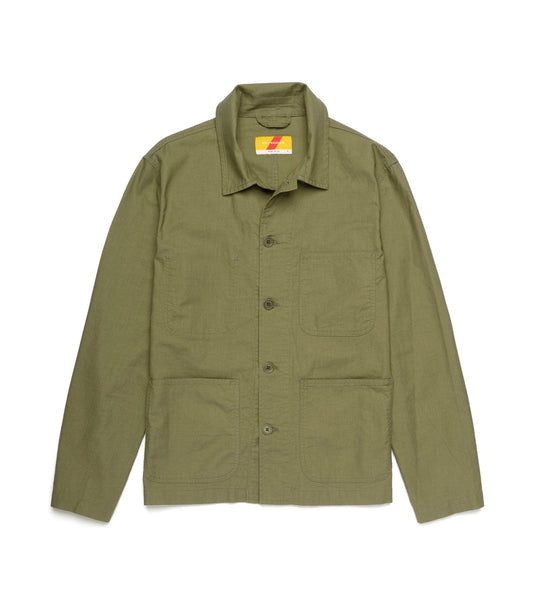 A man with light brown hair and a mustache stands indoors, wearing the Best Made Co. Panama Cloth Service Jacket over a tan shirt and blue jeans, looking at the camera while adjusting his jacket.