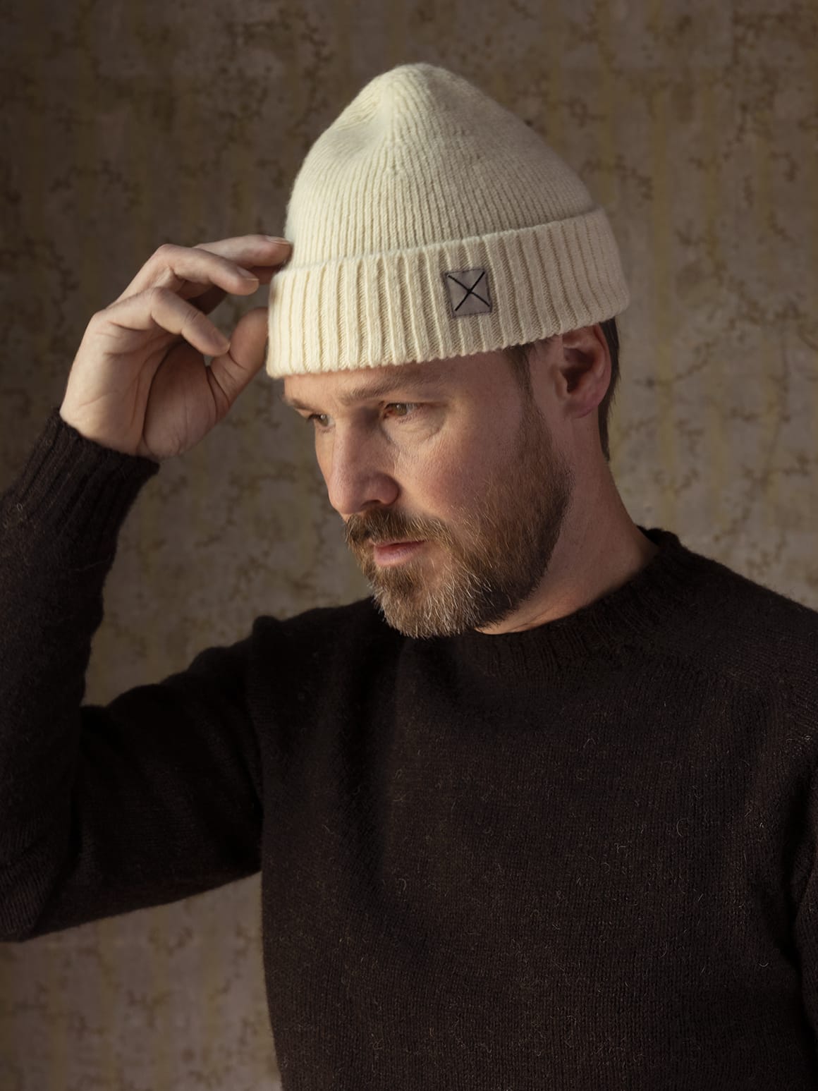 A man wearing a cream-colored, undyed Best Made Co. Shetland Cap of Courage, featuring a small square patch with an X on the folded cuff.