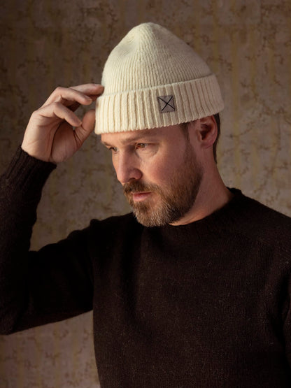 A man wearing a cream-colored, undyed Best Made Co. Shetland Cap of Courage, featuring a small square patch with an X on the folded cuff.