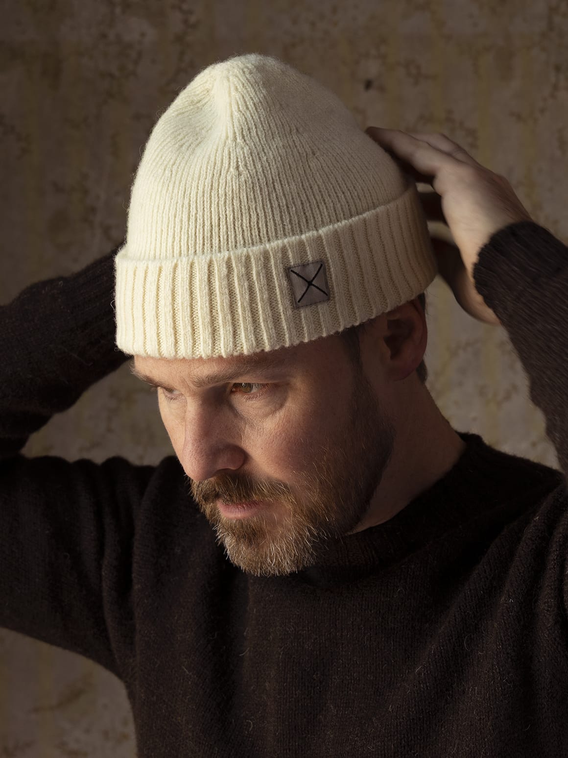 A person wearing the Best Made Co. Shetland Cap of Courage, a durable, cream-colored ribbed knit beanie made from undyed Shetland wool featuring a small square patch on the front cuff.