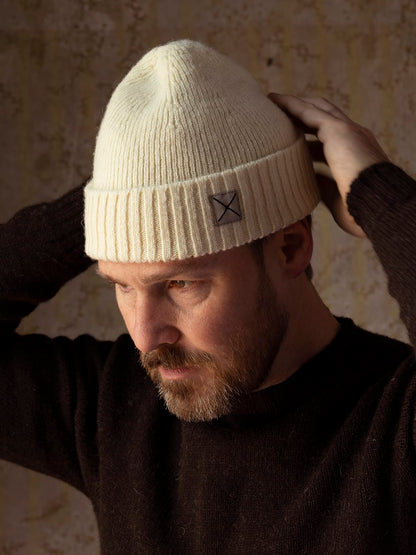 A person wearing the Best Made Co. Shetland Cap of Courage, a durable, cream-colored ribbed knit beanie made from undyed Shetland wool featuring a small square patch on the front cuff.