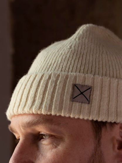 Best Made Co. Shetland Cap of Courage: a cream-colored ribbed knit beanie made of undyed Shetland wool, featuring a square patch with an X embroidery on the cuff.