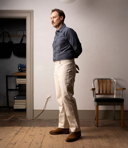 A man stands indoors on a wooden floor, facing left with hands behind his back, wearing a blue shirt, brown shoes, and Best Made Co. Standard 5-Pocket Pant M2 Undyed. Behind him are a wooden chair and a shelf with bags, books, and an old sewing machine.
