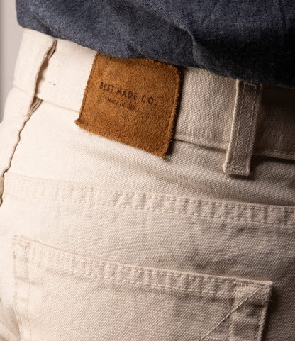 Close-up of a person wearing Best Made Co. Standard 5-Pocket Pant M2 Undyed—off-white, heavyweight cotton canvas jeans with a brown leather brand patch—paired with a tucked-in dark blue shirt.