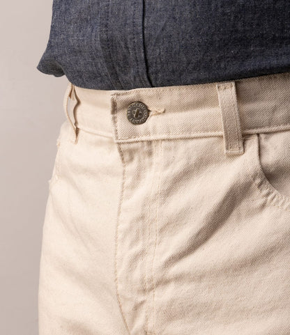Close-up of a person wearing Best Made Co. Standard 5-Pocket Pant M2 Undyed, heavyweight cotton canvas in cream with a visible metal button, paired with a tucked-in blue chambray shirt. Focus on the waist and upper pants.