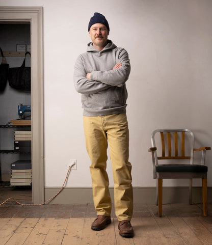 A man stands indoors with arms crossed, wearing a navy beanie, gray hoodie, Best Made Co. Standard 5-Pocket Pant M2 Khaki, and brown shoes. He is on a wooden floor near a chair; shelves and a sewing machine are in the background.