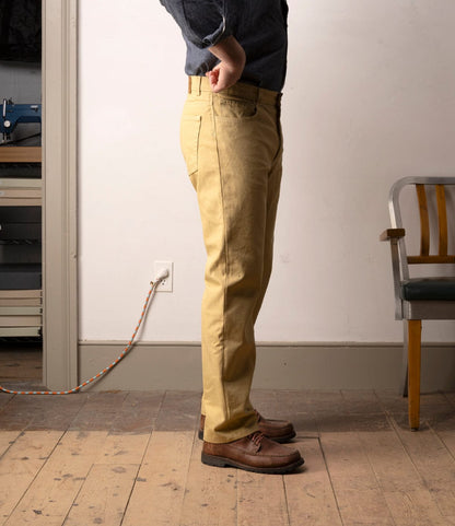 A person stands sideways indoors wearing a blue shirt, Best Made Co. Standard 5-Pocket Pant M2 Khaki, and brown shoes. The room features wooden floors, a chair, a shelf, and an orange extension cord plugged into a wall outlet.
